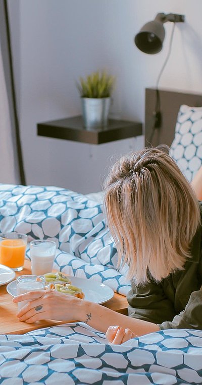 Pauschalen für Wochenendunterkünfte in Ostbayern - junge blonde Frau liegt im Bett neben einer Frühstücksplatte, gefüllt mit O-Saft, Milch und belegten Brötchen