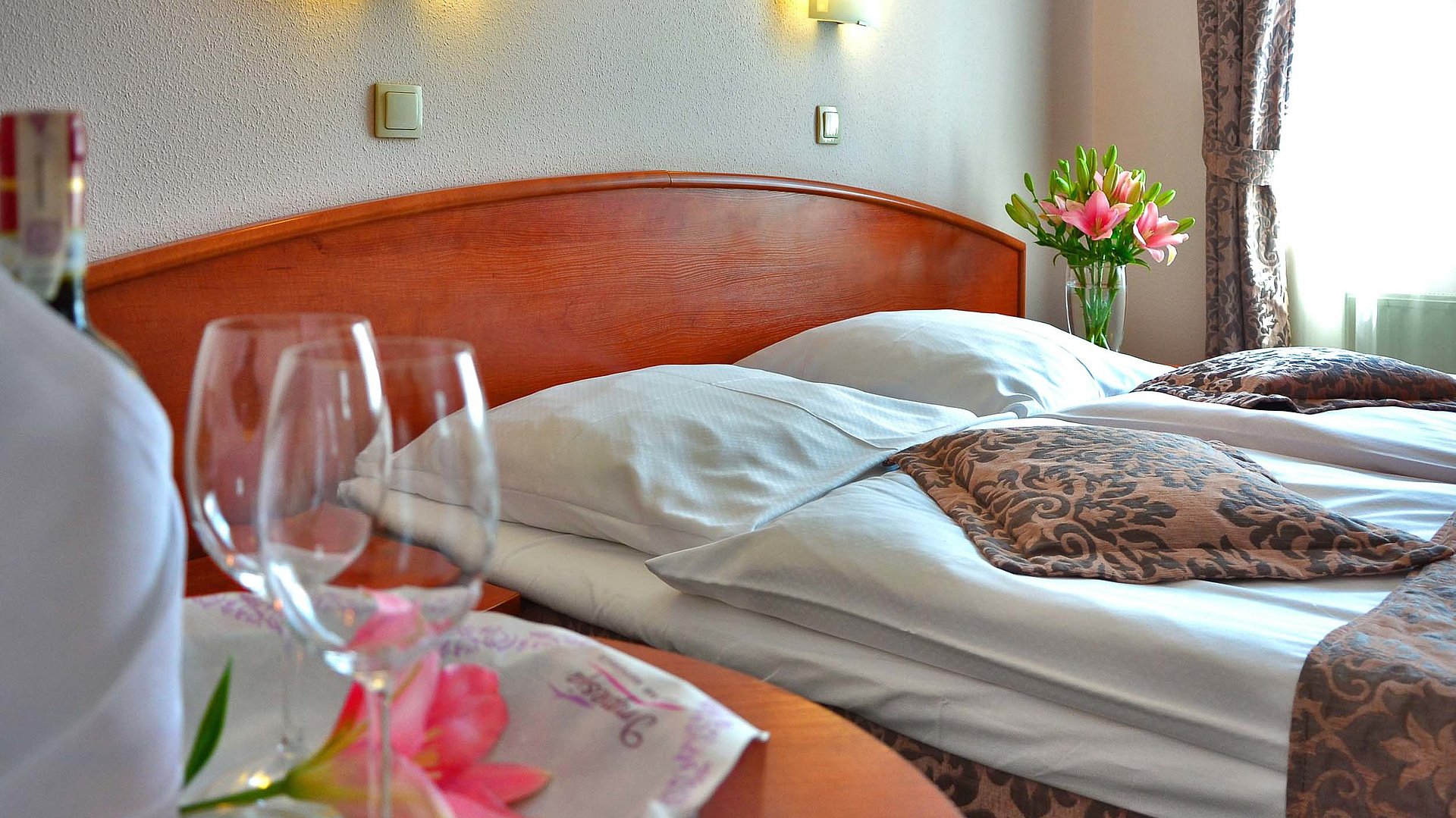 Pauschalangebote während der Woche für Unterkünfte in der Fränkischen Schweiz - Fokus auf sehr ordentliches Hotelbett; zwei Weingläser im Vordergrund und Blumen auf dem Nachttisch im Hintergrund 