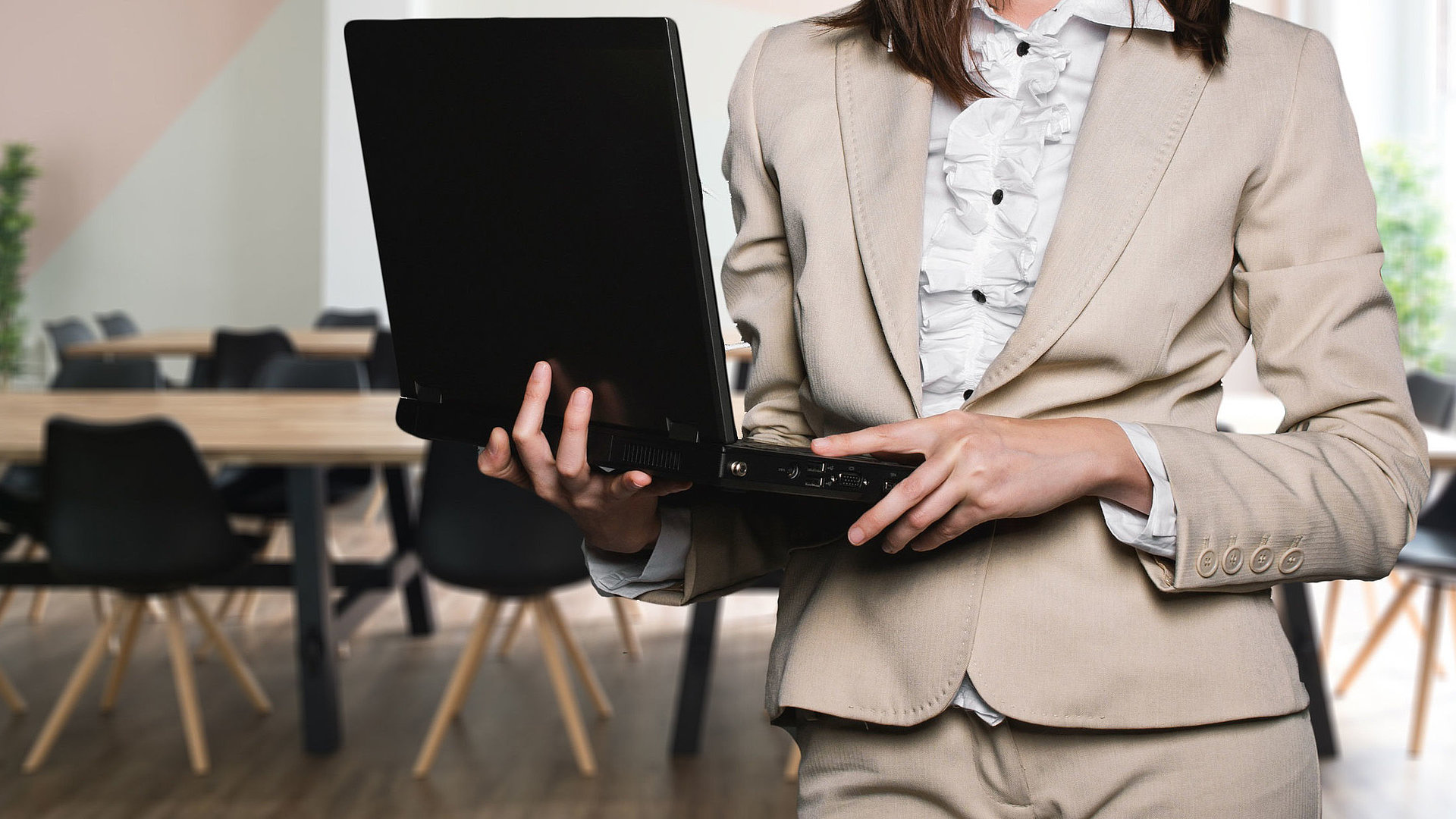Unterkünfte für Geschäftsreisende in Nürnberg - Geschäftsfrau im beigen Anzug, weißer Bluse und schwarzem Laptop auf dem Arm steht in vor einem Meeting-Tisch