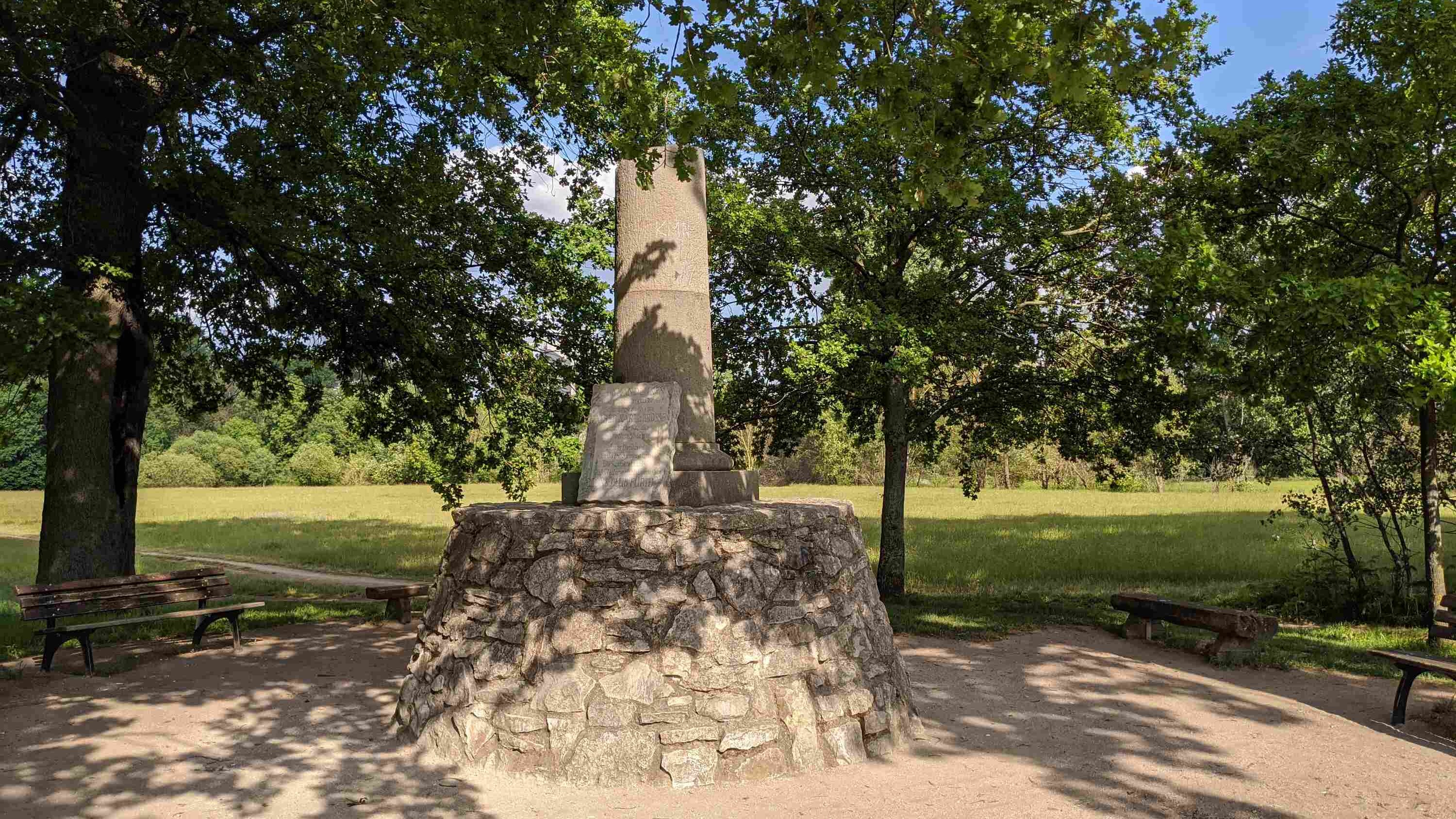 Gedenksäule mit Tafel unter schattigen Bäumen
