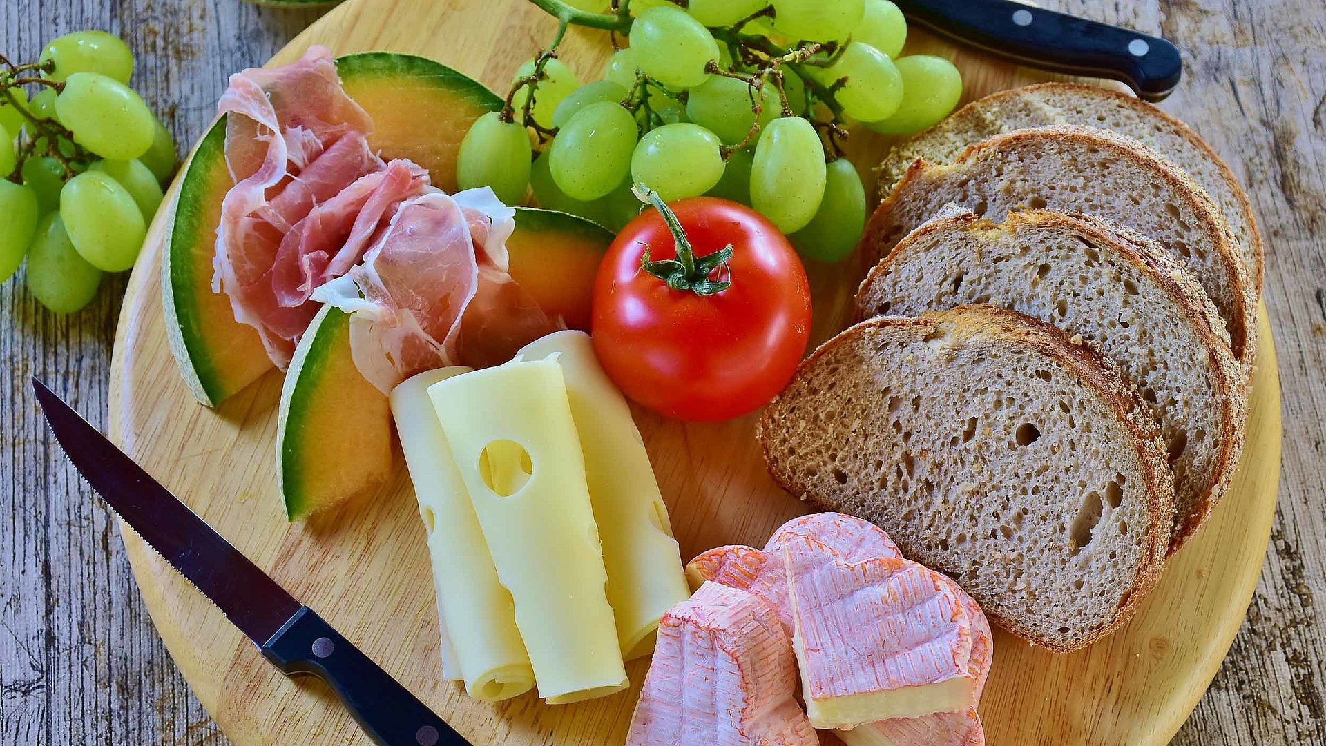 Brotzeit in Ostbayern - rundes Holzbrett mit Brot, französischem Käse, Scheibenkäse, Tomate und Weintrauben 