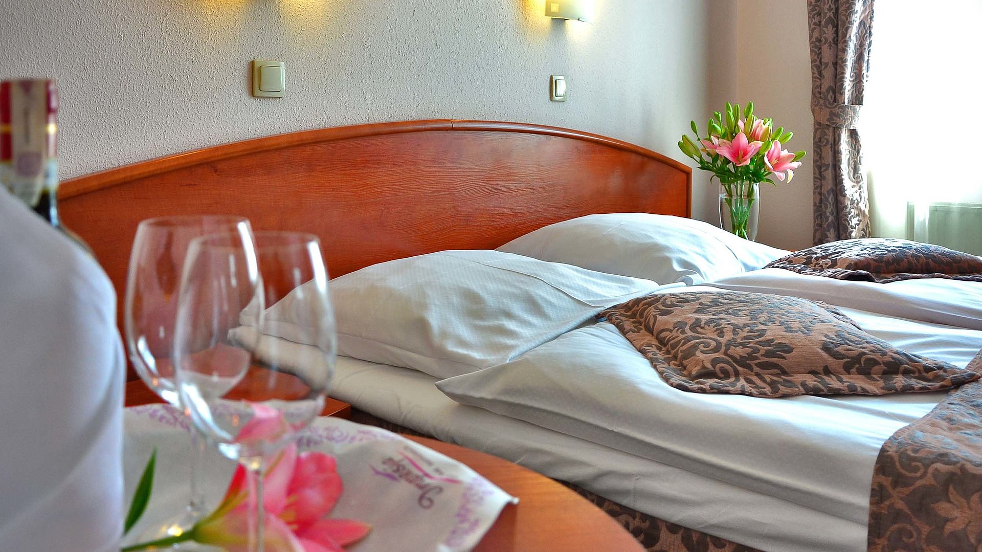 Pauschalangebot für eine Woche in Kempten im Allgäu - im Hotelzimmer von Gardinen durchleuchtet; Weingläser auf Tisch und Blumenstrauß auf Nachttisch