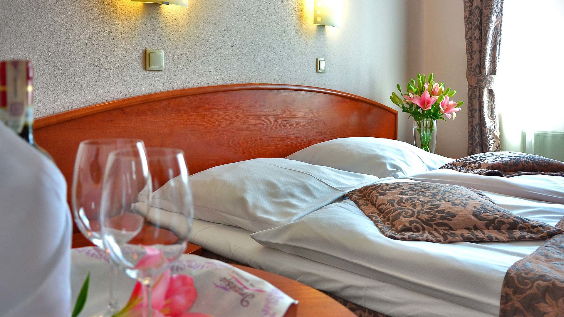 Pauschalangebote für eine Woche in Mittelfranken - im Hotelzimmer von Gardinen durchleuchtet; Weingläser auf Tisch und Blumenstrauß auf Nachttisch