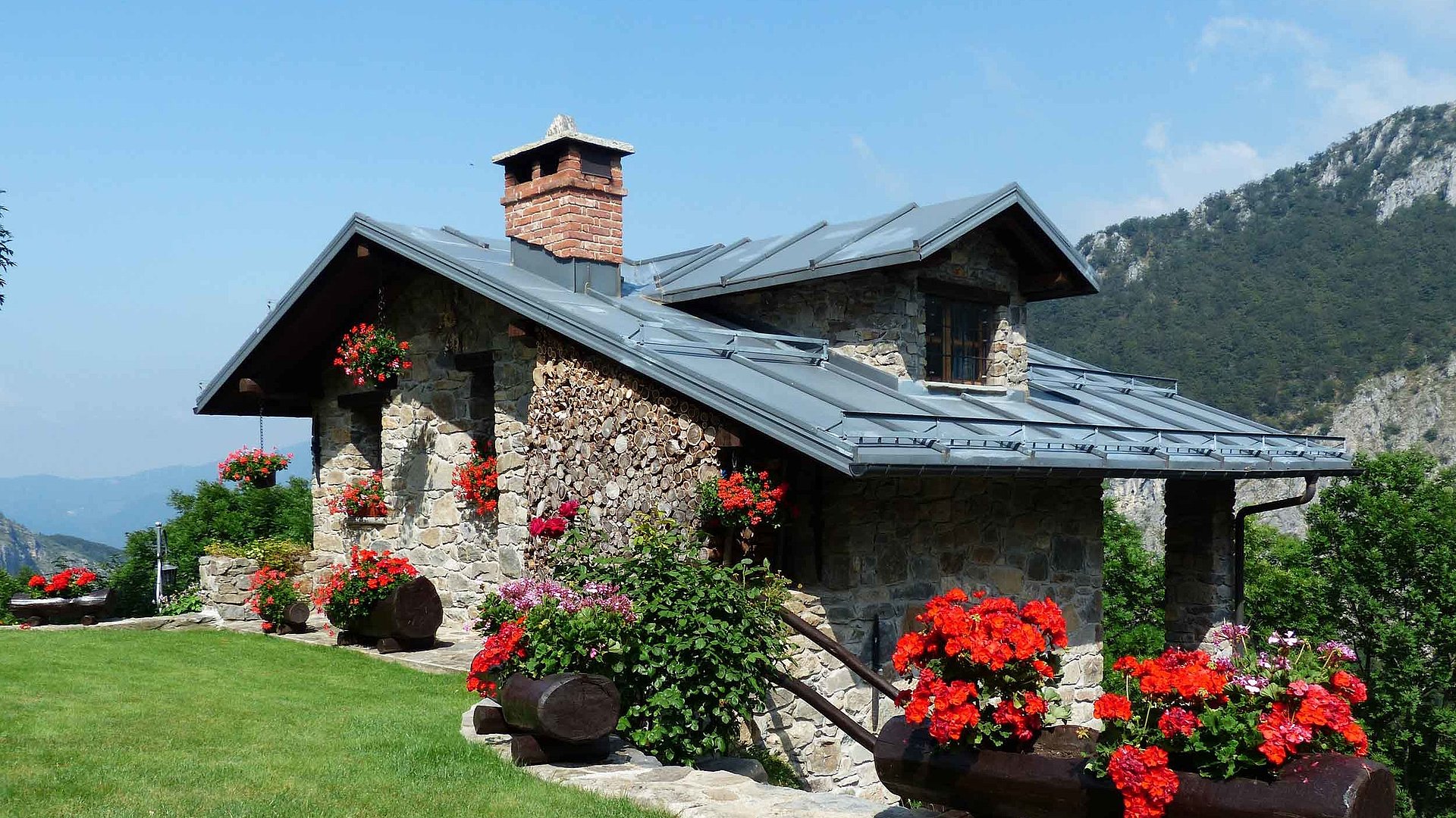 Ferienwohnungen und Ferienhäuser für Gruppen im Oberallgäu - mittelgroßes Ferienhaus mit grüner Wiese und Blumenbeeten, im Hintergrund Hügellandschaft bei klaren Himmel