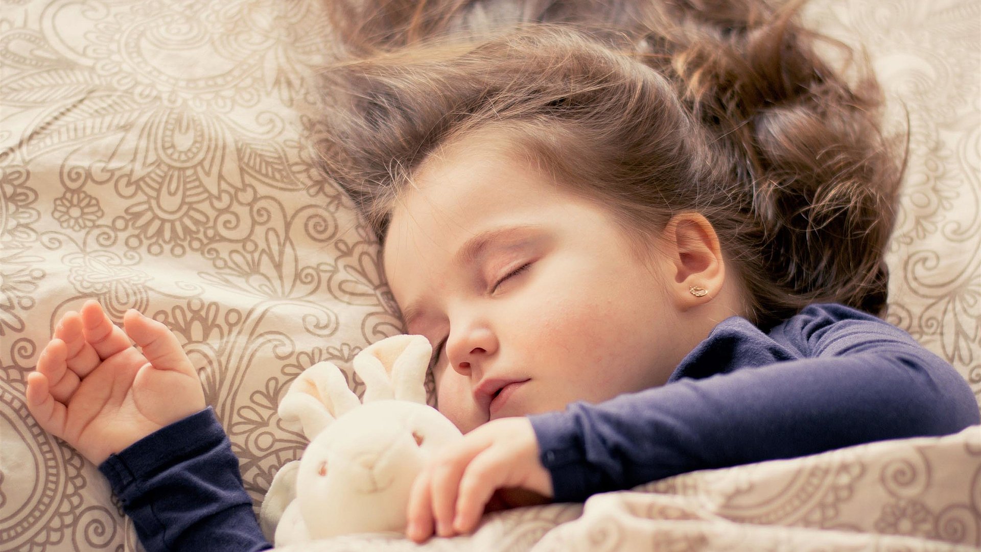 Kinderfreundliche Unterkünfte in Oberbayern - kleines Mädchen mit Plüschtierhäschen liegt schlafend im Bett  