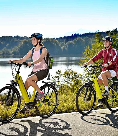 E-Bike im Fichtelgebirge - zwei Radfahrer mit E-Bike fahren auf Radweg bei sonnigem Wetter an einem See vorbei; Waldlandschaft im Hintergrund