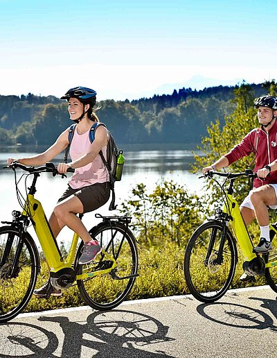 Radfahren und Radwandern in Oberbayern - zwei Radfahrer mit E-Bike fahren auf Radweg bei sonnigem Wetter an einem See vorbei; Waldlandschaft im Hintergrund