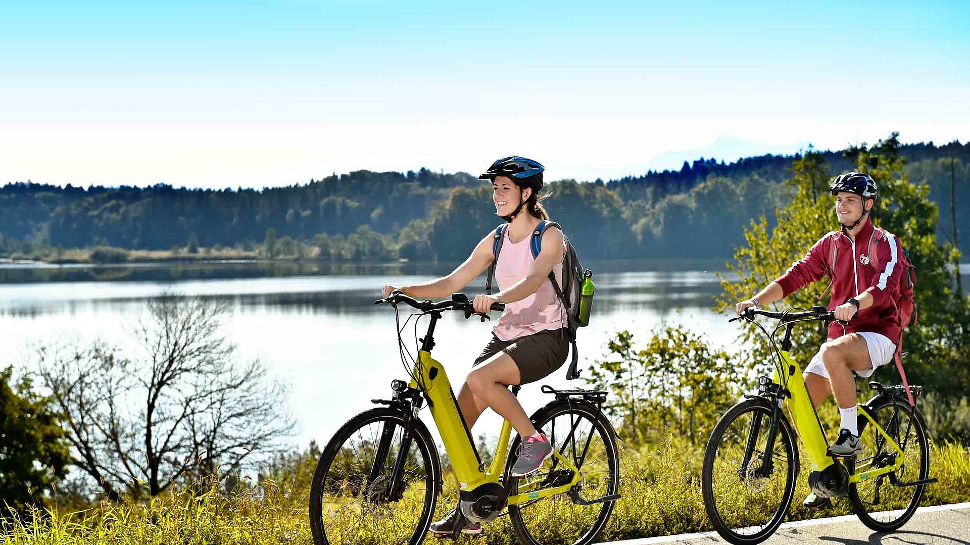 Aktivurlaub mit dem Rad im Fränkischen Seenland - zwei Radfahrer mit E-Bike fahren auf Radweg bei sonnigem Wetter an einem See vorbei; Waldlandschaft im Hintergrund