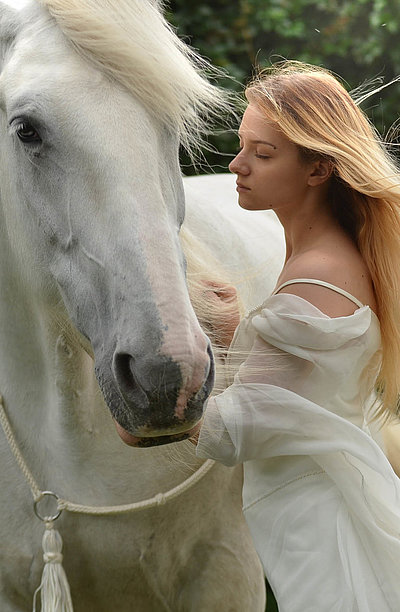 Urlaub auf dem Bauernhof - junge Frau mit langem blonden Haar und weißem Kleid streichelt weißes Pferd