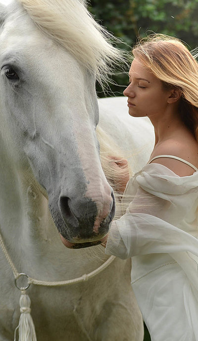 Urlaub auf dem Bauernhof in Oberfranken - junge Frau mit langem blonden Haar und weißem Kleid streichelt weißes Pferd
