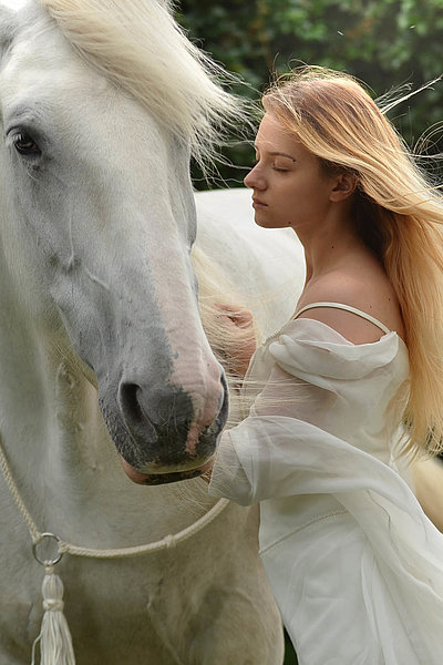 Urlaub auf dem Bauernhof in Oberbayern - junge Frau mit langem blonden Haar und weißem Kleid streichelt weißes Pferd