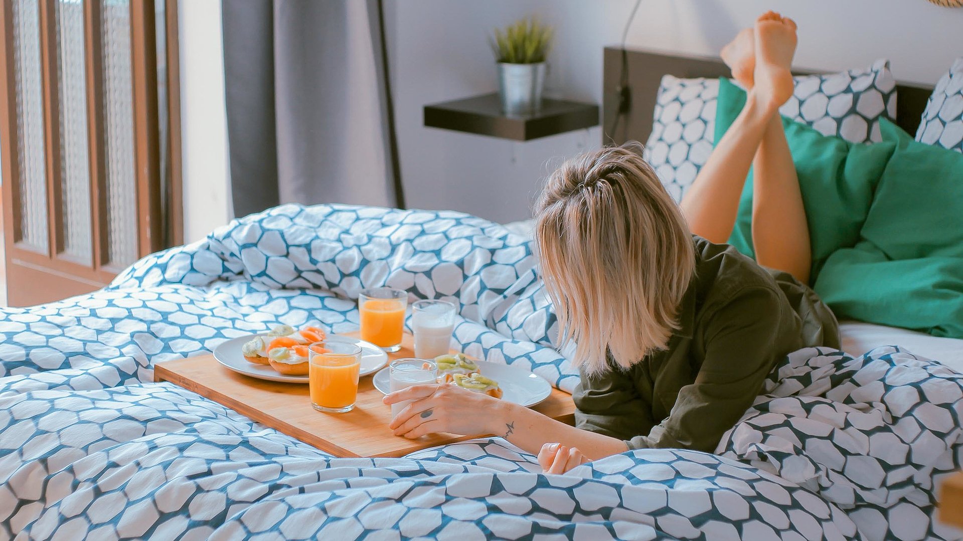 Hotels in Nürnberg - junge Frau liegt auf dem Bett neben einer vollen Frühstücksplatte, mit O-Saft, belegten Brötchen und Milch