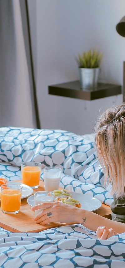 Pauschalen für Wochenenden in Unterfranken - junge blonde Frau liegt im Bett neben einer Frühstücksplatte, gefüllt mit O-Saft, Milch und belegten Brötchen
