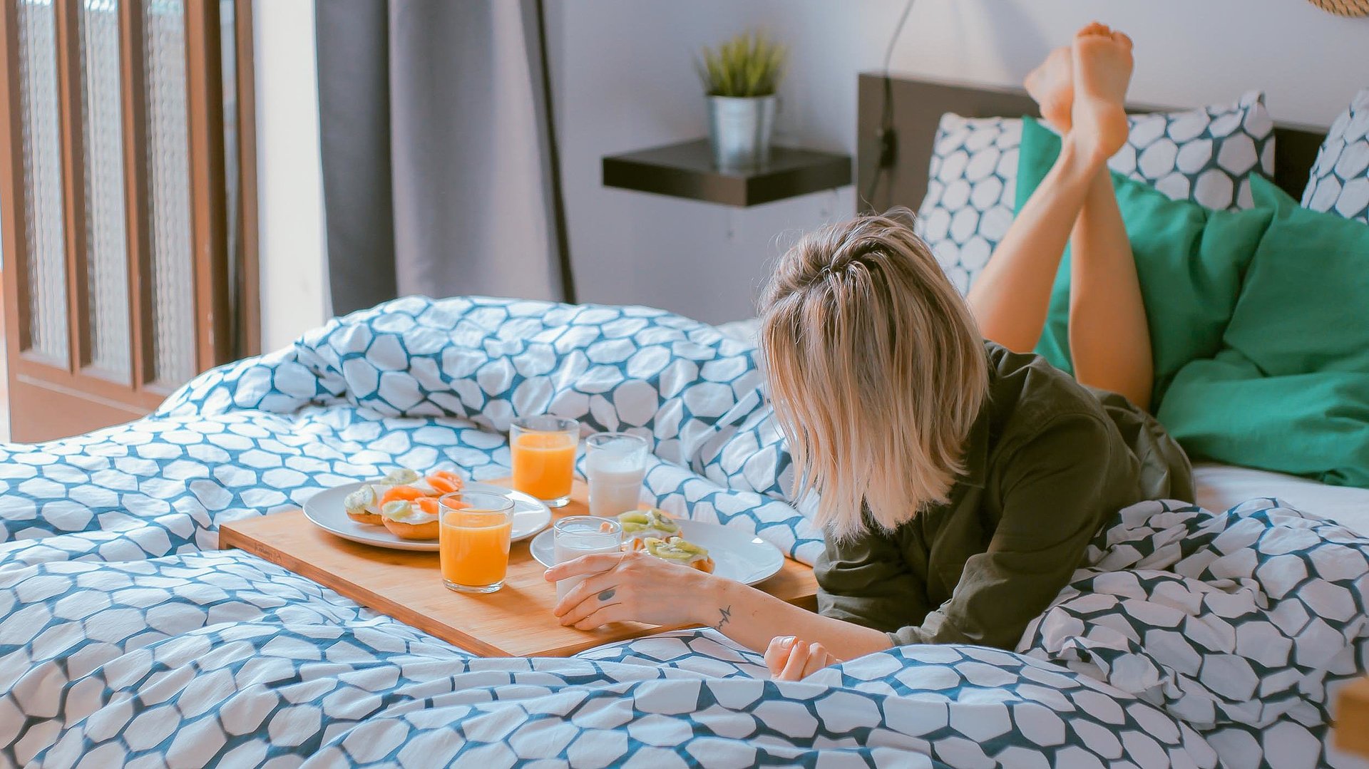 Pauschalen für eine Woche in Oberbayern - junge blonde Frau liegt im Bett neben einer Frühstücksplatte, gefüllt mit O-Saft, Milch und belegten Brötchen