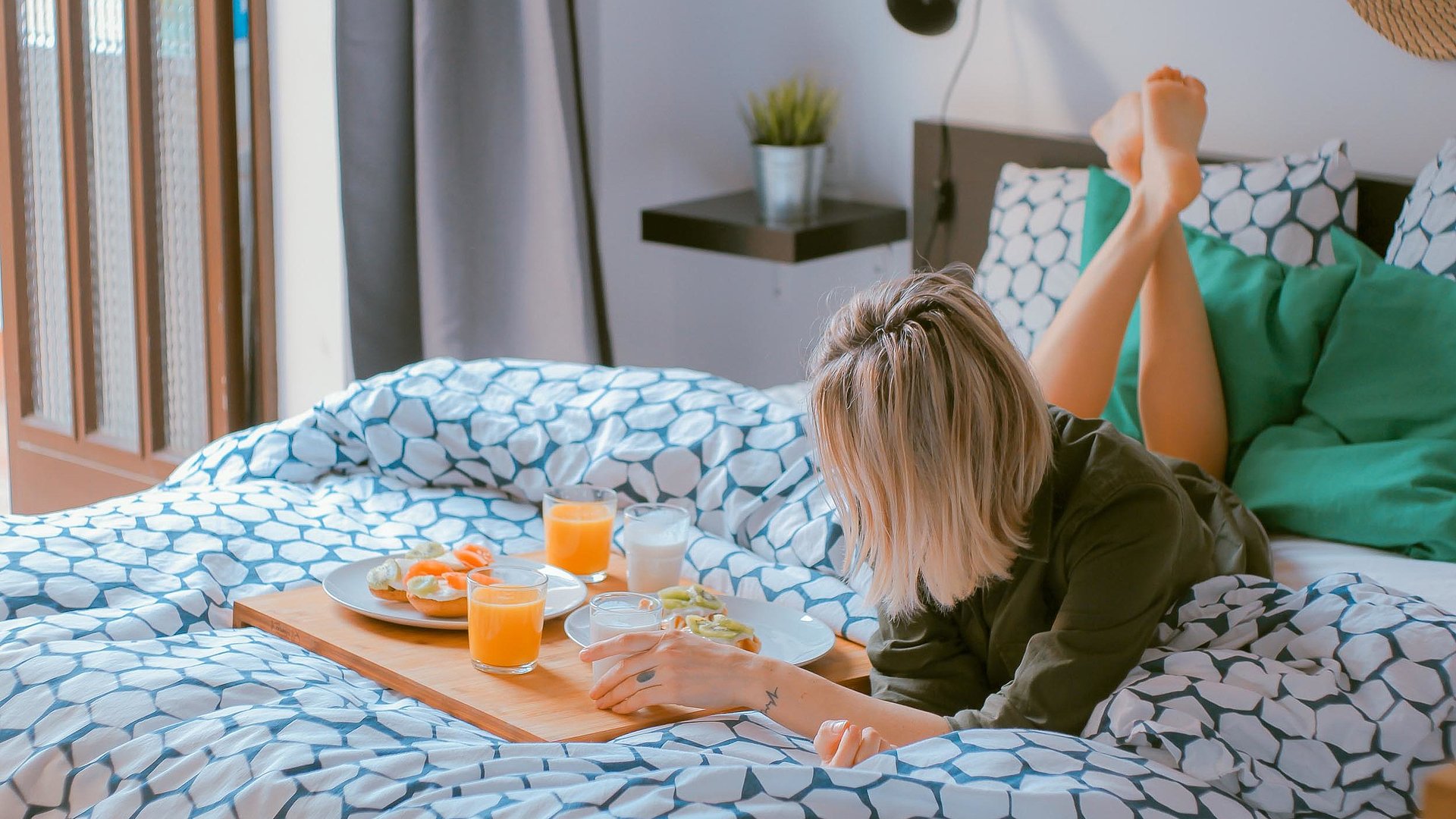 Pauschalangebote für Wochentagsunterkünfte in Unterfranken - junge blonde Frau liegt im Bett neben einer Frühstücksplatte, gefüllt mit O-Saft, Milch und belegten Brötchen 