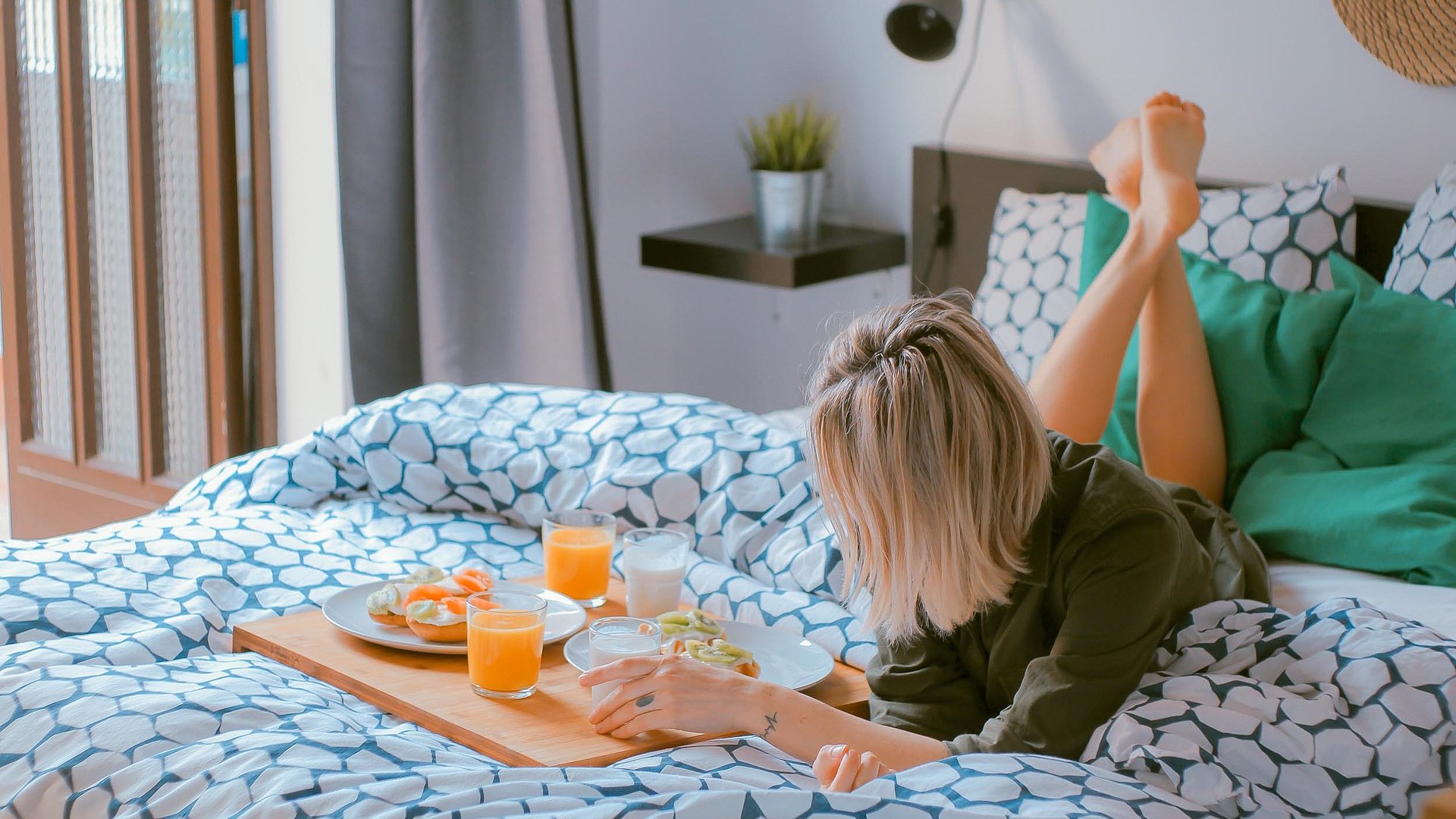 Pauschalangebote für Unterkünfte zu Wochentagen im Oberallgäu - junge blonde Frau liegt im Bett, vor Ihr eine volle Frühstücksplatte mit O-Saft, Milch und belegten Brötchen