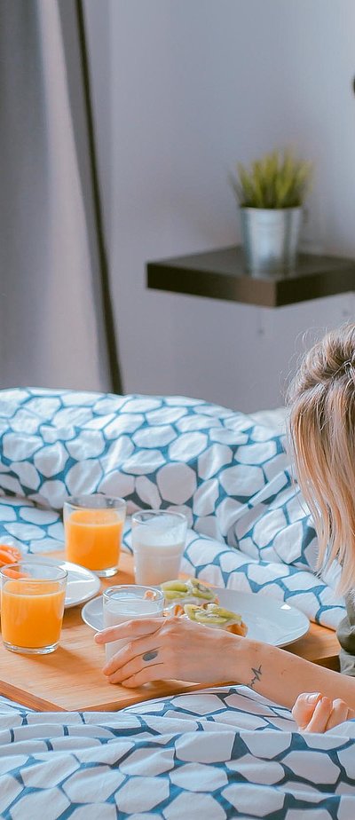 Pauschalangebote Unterkunft in Mittelfranken - junge blonde Frau liegt im Bett neben einer Frühstücksplatte, gefüllt mit O-Saft, Milch und belegten Brötchen 
