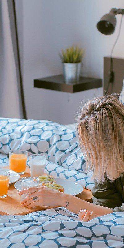 Pauschalangebote für Unterkünfte zu Wochentagen im Oberallgäu - junge blonde Frau liegt im Bett neben einer Frühstücksplatte, gefüllt mit O-Saft, Milch und belegten Brötchen