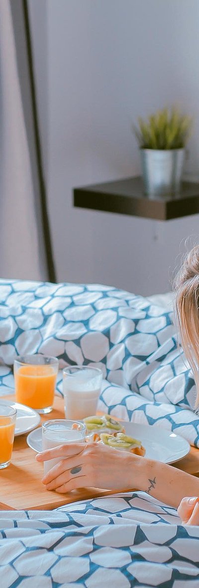 Ferienwohnungen und Appartements für Gruppen in Oberfranken - junge blonde Frau liegt im Bett neben einer Frühstücksplatte, gefüllt mit O-Saft, Milch und belegten Brötchen 