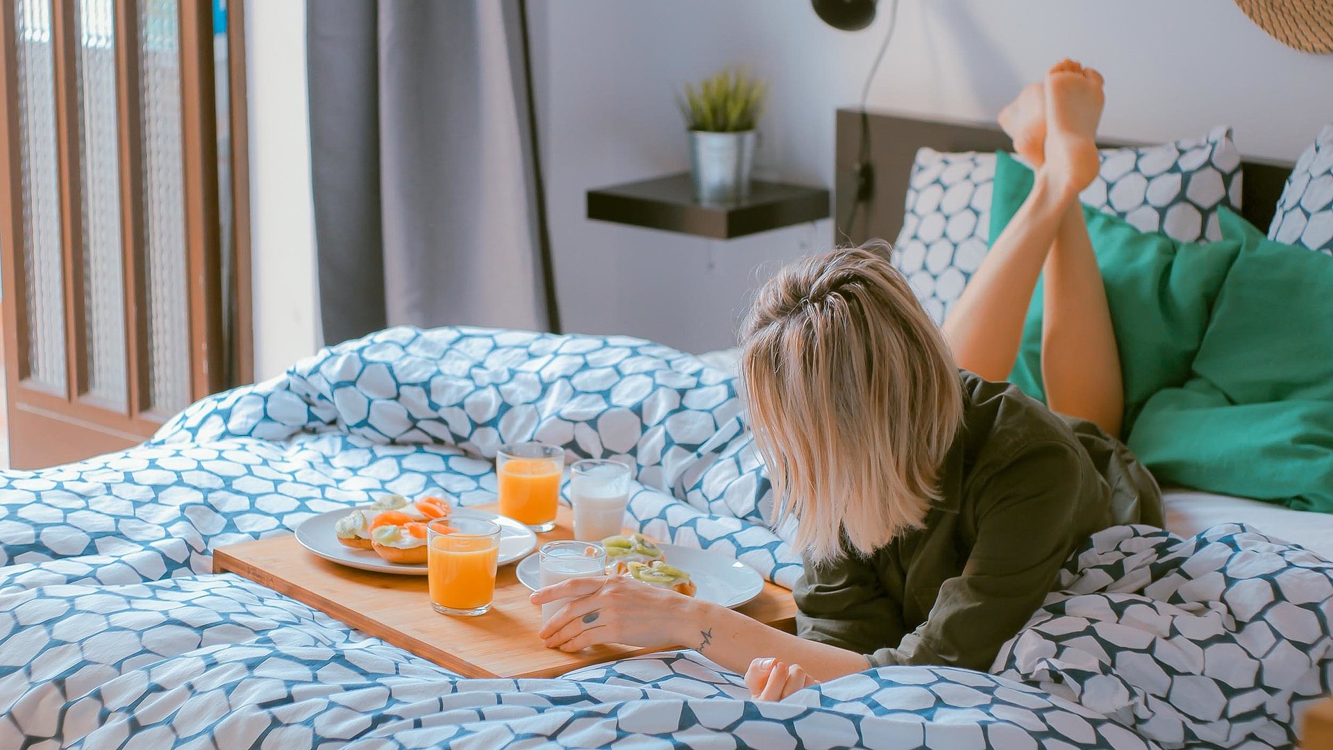 Pauschalangebote für Wochentagsunterkünfte in Oberfranken - junge blonde Frau liegt im Bett neben einer Frühstücksplatte, gefüllt mit O-Saft, Milch und belegten Brötchen 