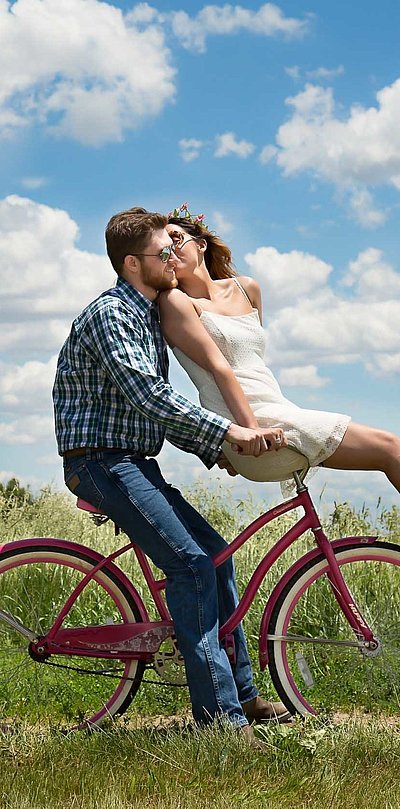 Radfahren und Radwandern in Ostbayern - junger Mann sitzt auf Fahrrad, seine Partnerin ist auf dem vorderen Gepäckträger und küsst ihn; sonniges Wetter auf einem Feld