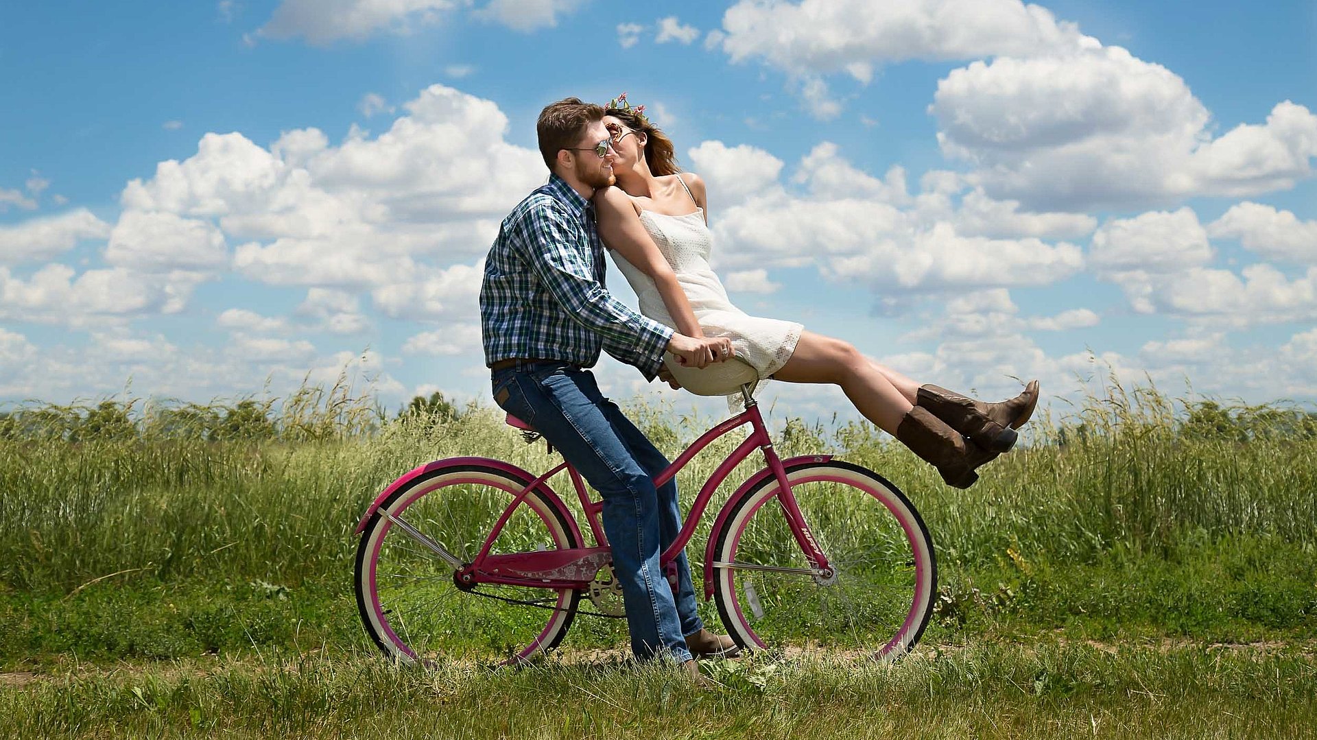 Radfahren und Radwandern in Ostbayern - junger Mann sitzt auf Fahrrad, seine Partnerin ist auf dem vorderen Gepäckträger und küsst ihn; sonniges Wetter auf einem Feld 