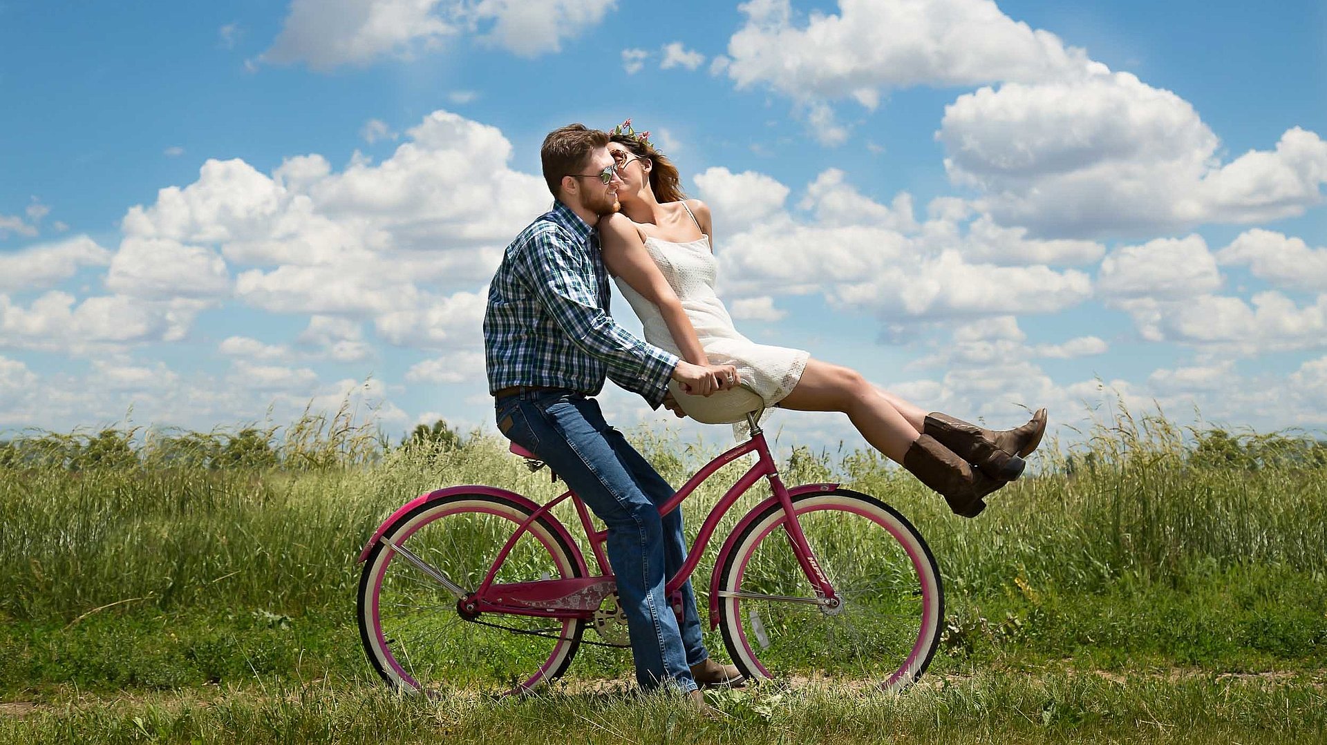 Radfahren und Radwandern in Oberbayern - junger Mann sitzt auf Fahrrad, seine Partnerin ist auf dem vorderen Gepäckträger und küsst ihn; sonniges Wetter auf einem Feld