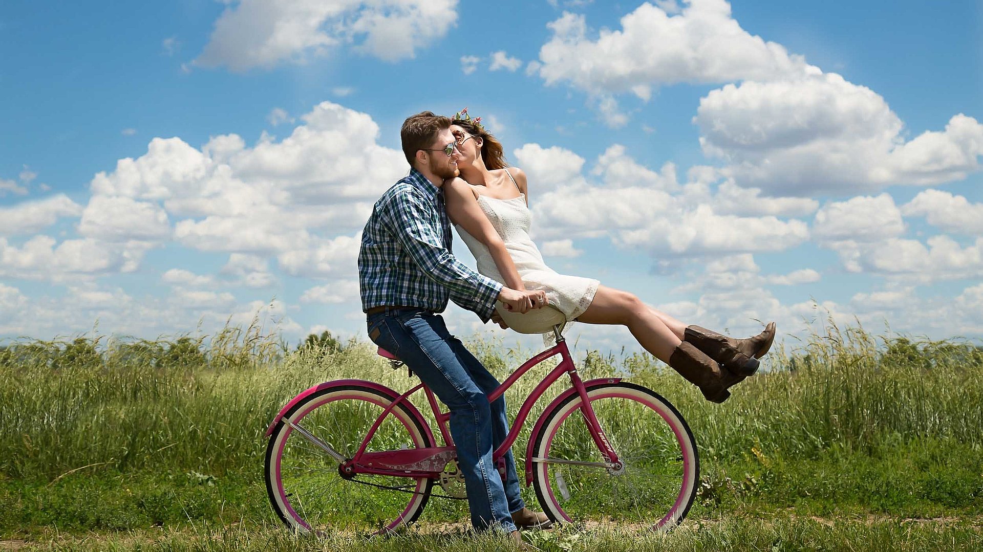 Radfahren und Radwandern in Oberfranken - junger Mann sitzt auf Fahrrad, seine Partnerin ist auf dem vorderen Gepäckträger und küsst ihn; sonniges Wetter auf einem Feld