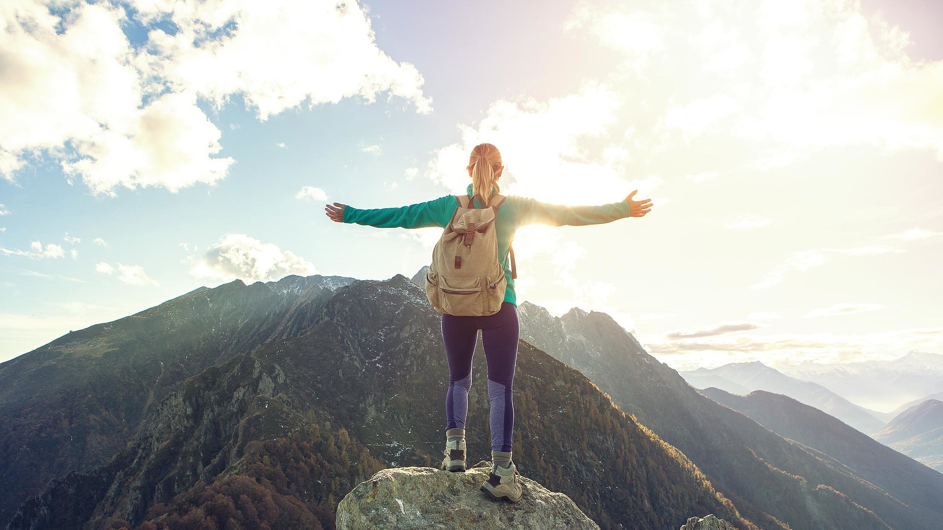 Outdoor-Freizeitangebote im Oberallgäu -junge Frau mit Wanderrucksack, auf einem Gipfel, mit Rücken zur Kamera streckt Arme Richtung Berge aus